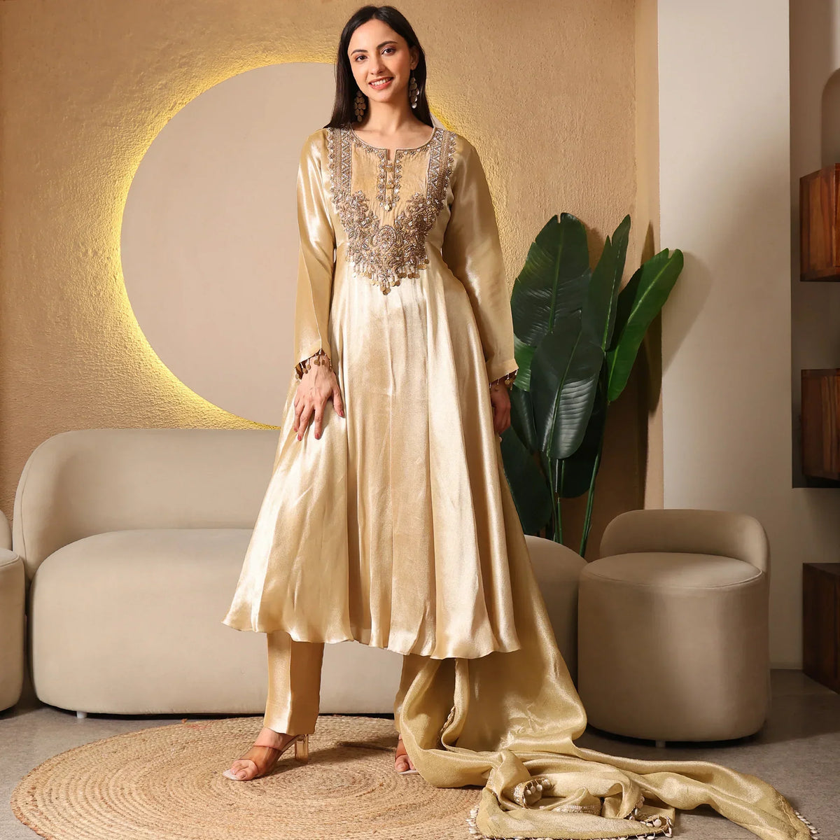 Woman in gold Banarasi Anarkali suit with embroidery, standing in stylish living room