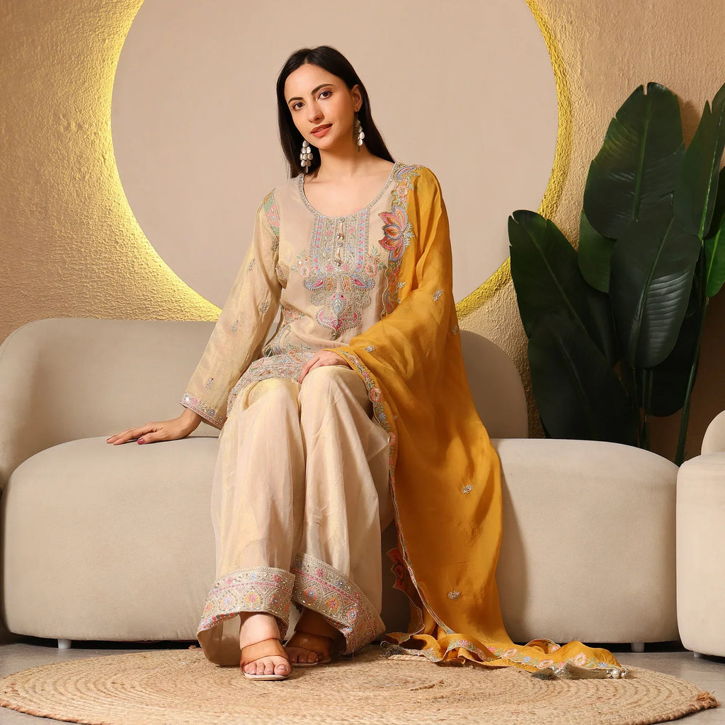 Woman in beige and yellow ethnic Anarkali suit with embroidery, sitting on a beige sofa.