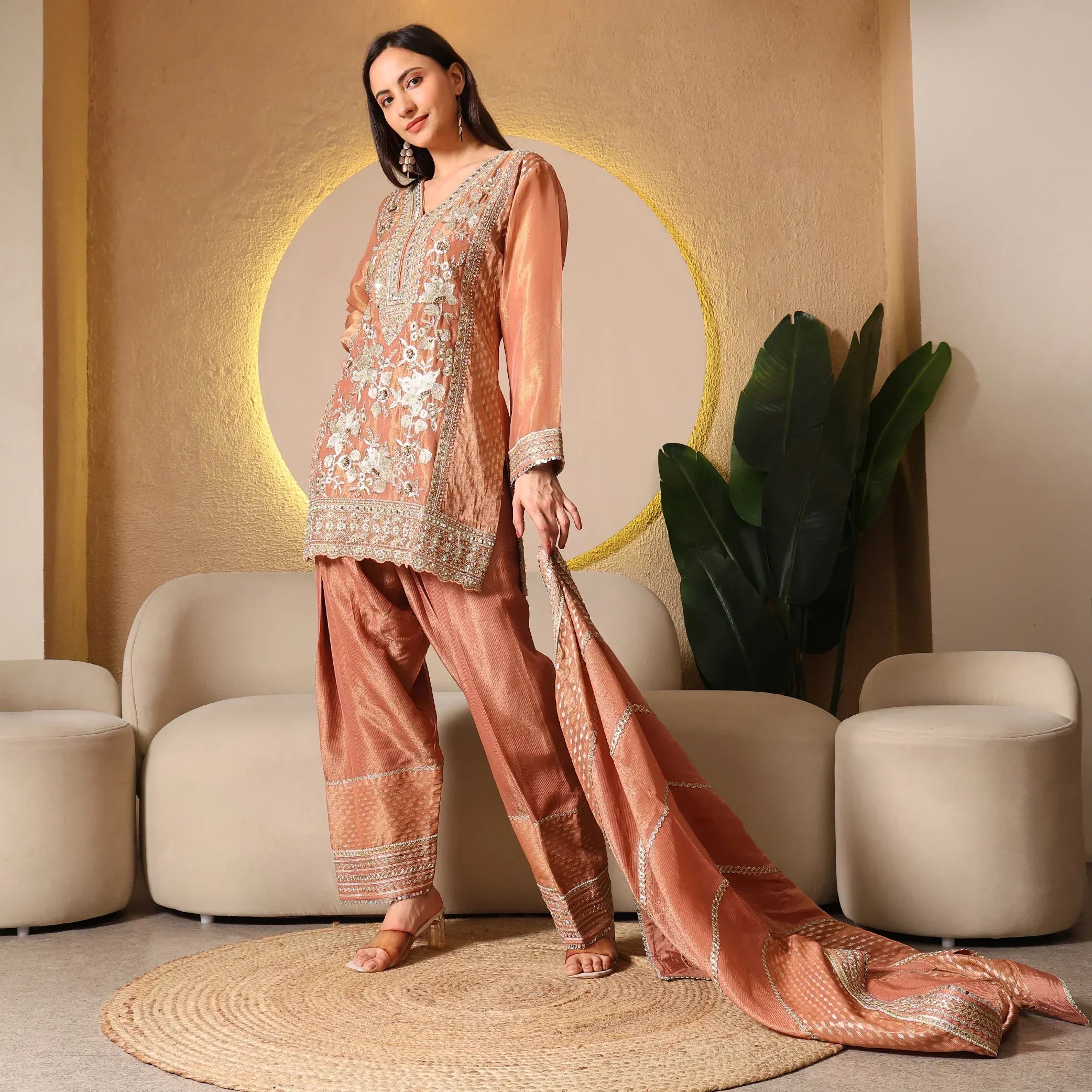 Woman modeling an embroidered peach ethnic suit with dupatta, seated indoor setting