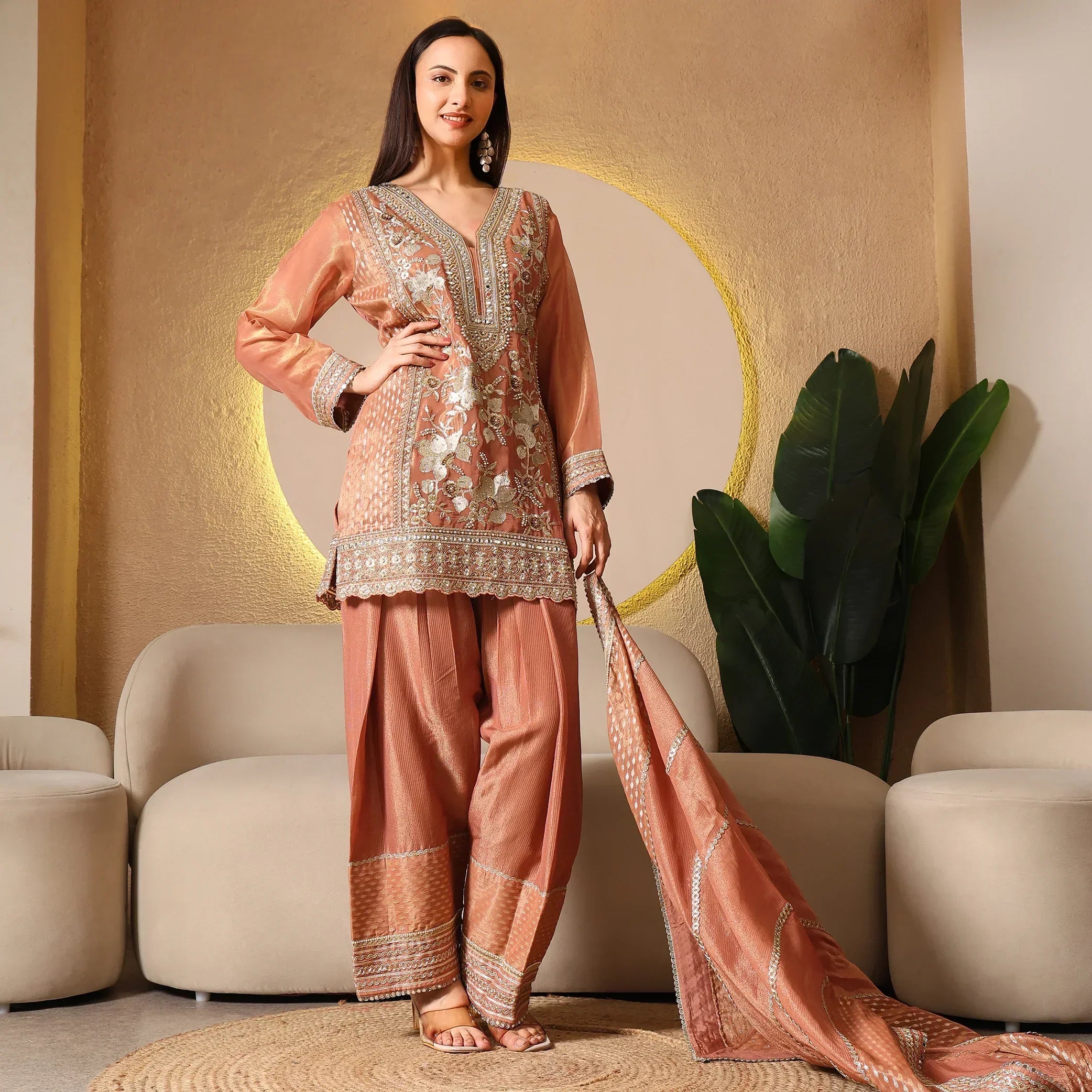 Woman wearing a peach embroidered ethnic suit with dupatta, standing in a modern living room