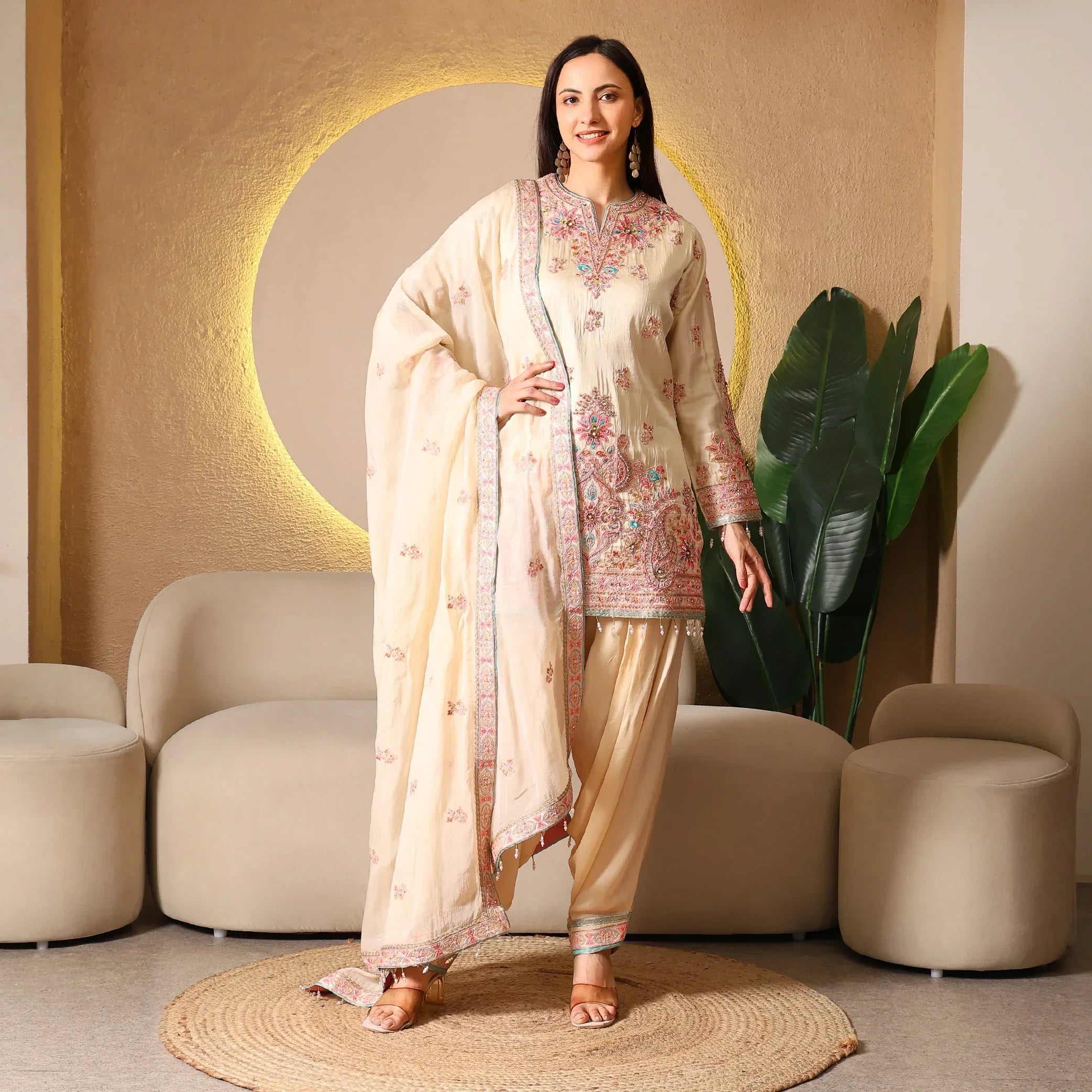 Woman in a cream embroidered ethnic suit with dupatta, posing in a modern living room.