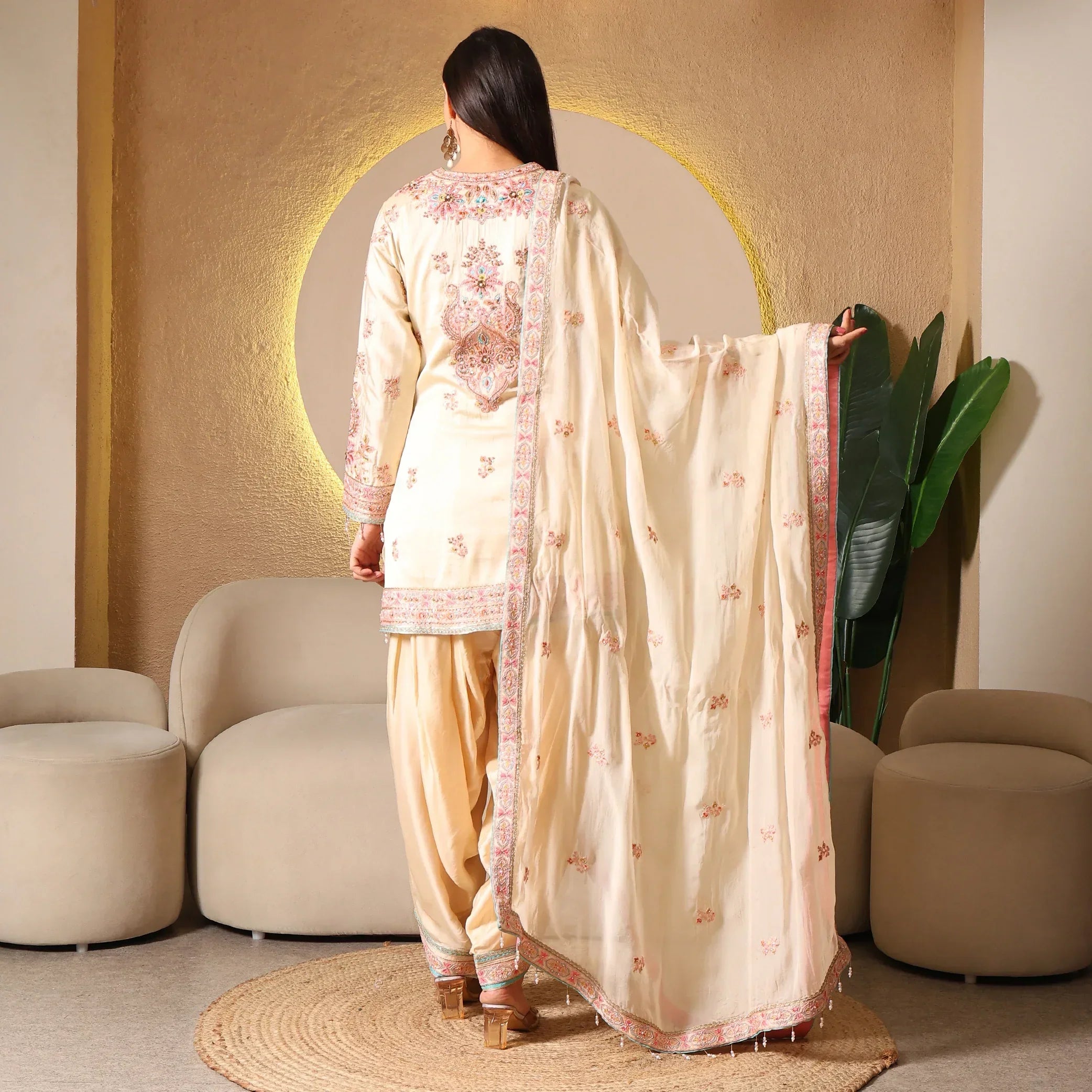 Woman in cream embroidered ethnic suit with matching dupatta, standing in modern living room