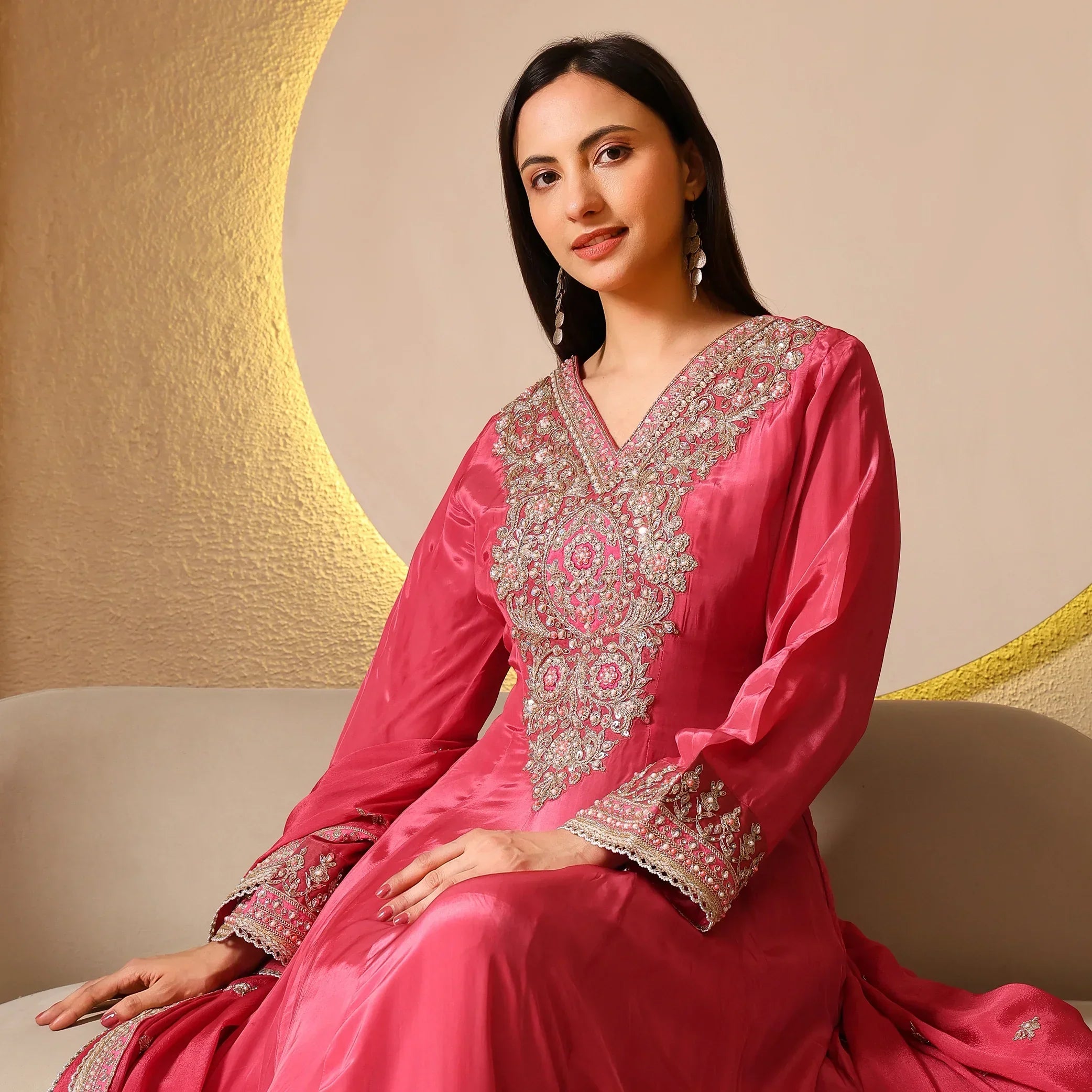 Woman in pink embellished Anarkali suit with intricate embroidery, seated on beige sofa indoors
