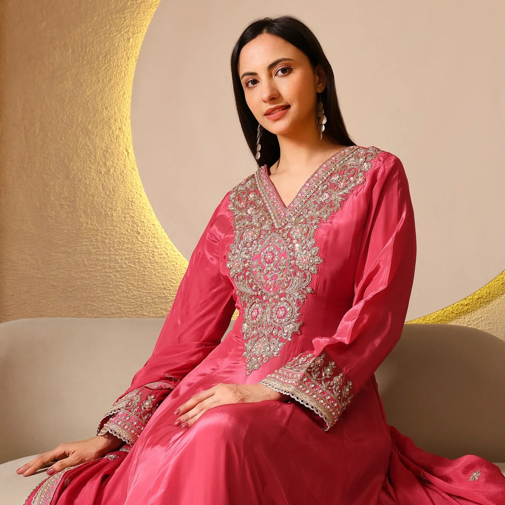 Woman in pink embellished Anarkali suit with intricate embroidery, seated on beige sofa indoors