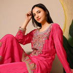 Woman wearing a bright pink embroidered ethnic suit with gold detailing, sitting indoors