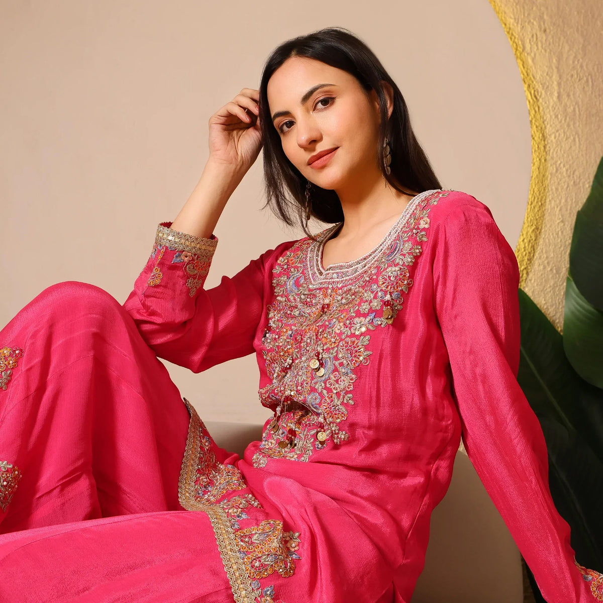 Woman wearing a bright pink embroidered ethnic suit with gold detailing, sitting indoors