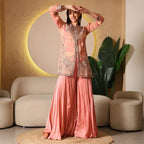 Woman in peach embroidered ethnic wear palazzo set, standing indoors by beige sofa and plants