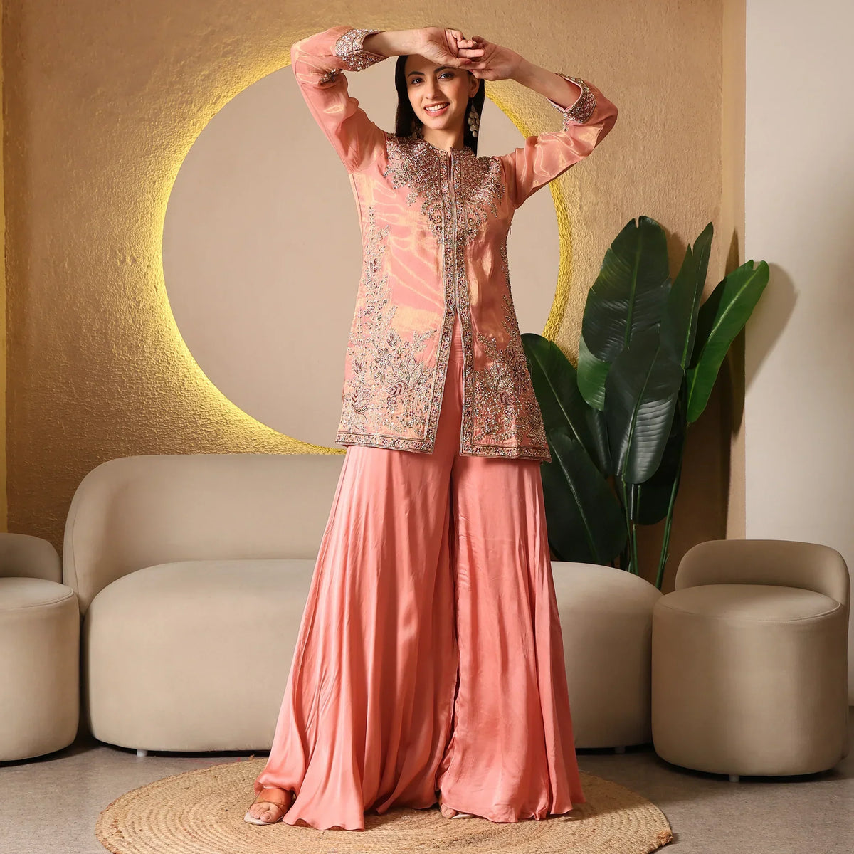 Woman in peach embroidered ethnic wear palazzo set, standing indoors by beige sofa and plants
