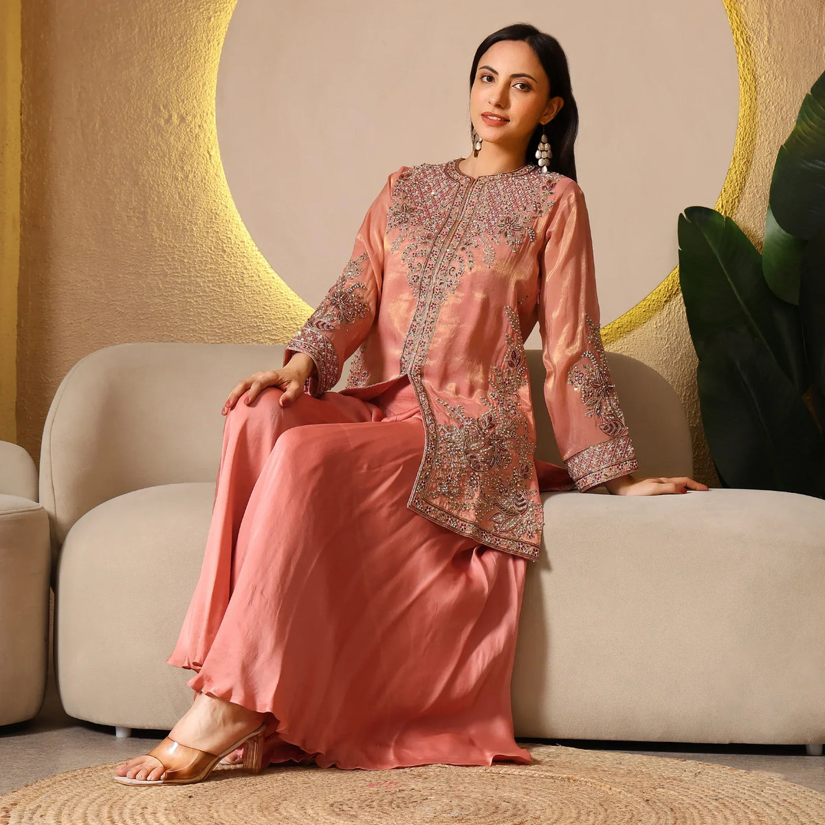 Woman in peach embroidered ethnic suit seated on beige sofa, festive outfit with heels