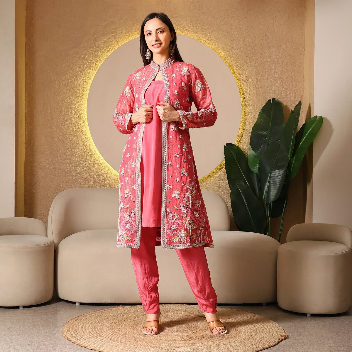 Woman in pink embroidered Banarasi ethnic suit with long jacket, standing in modern living room