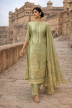 Woman in sage green Banarasi Anarkali suit with gold embroidery, walking by historic fort.