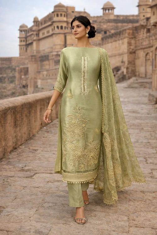 Woman in sage green Banarasi Anarkali suit with gold embroidery, walking by historic fort.