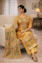 Woman in gold embroidered ethnic suit set with floral dupatta, sitting on sofa indoors