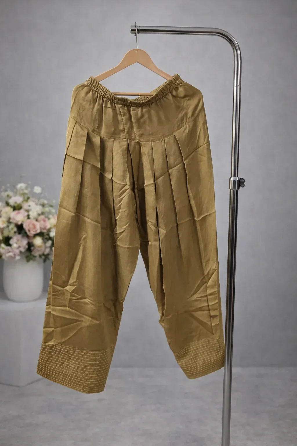 Gold pleated ethnic pants with elastic waistband, displayed on hanger, floral decor in background