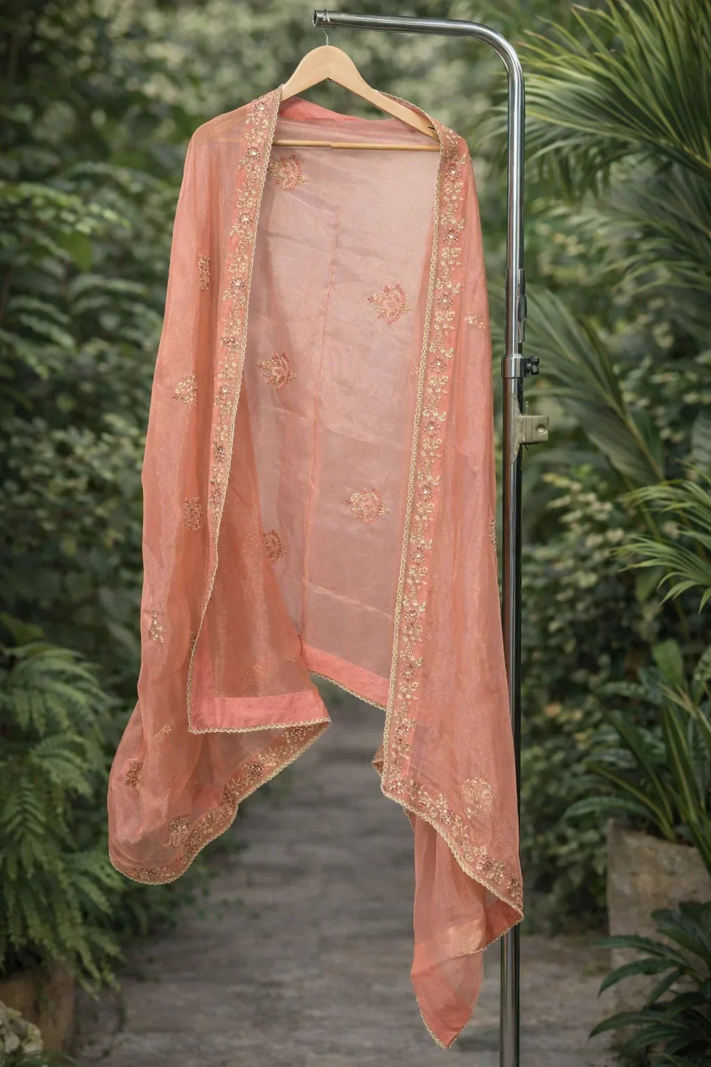 Peach embroidered dupatta with gold border displayed on hanger, outdoor greenery background
