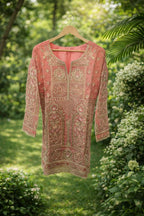 Pink embroidered ethnic kurta with gold thread work, displayed outdoors on a hanger.