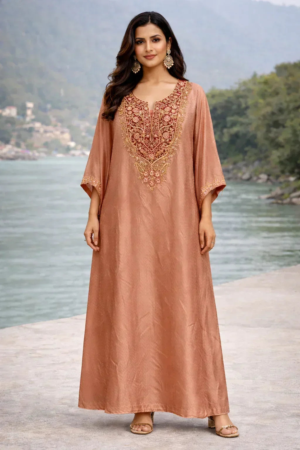 Woman in rose gold ethnic Anarkali dress with ornate embroidery, standing outdoors by a river