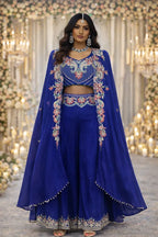 Woman in a royal blue embroidered ethnic suit with floral patterns, jewelry, and flowing cape, standing in a decorated festive setting.